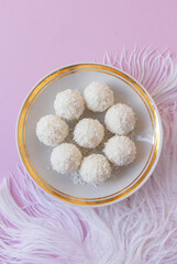 Top view of White Chocolate Candy With Coconut Topping On White Feather and pink background, sweet balls on plate flat lay with copy space. Romantic gift for celebration or anniversary