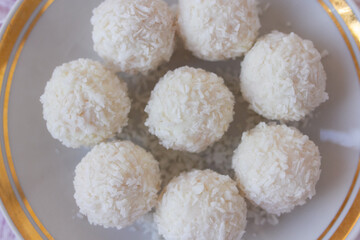 Top view of White Chocolate Candy With Coconut Topping On White Feather and pink background, sweet balls on plate close up flat lay with copy space. Romantic gift for celebration or anniversary