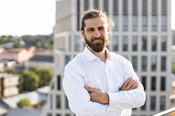 Confident businessman with beard and long hair standing on rooftop, wearing white shirt, arms...