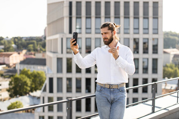 Businessman standing on rooftop in casual attire with smartphone taking selfie with cityscape in background. Represents modern business lifestyle urban life technology use remote work social media.