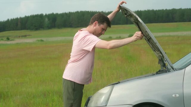 Young man opens car hood and inspects engine for signs of damage. Male lifts bonnet of automobile and tries to identify source of strange sounds