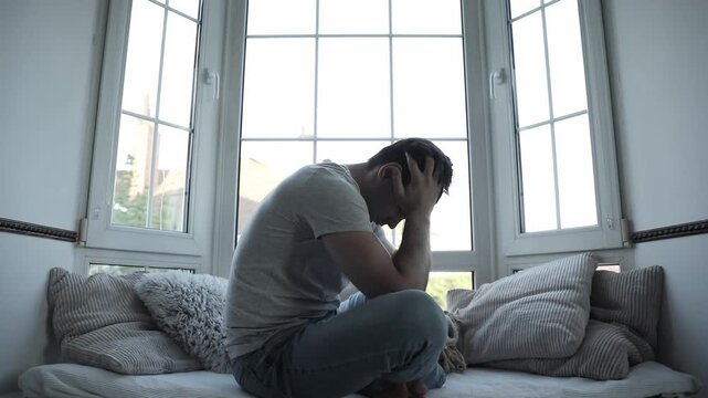 Depressed, sad, frustrated, crying, scared, instability mental and bipolar young man sitting on the windowsill at home. Headache because overworked and stress