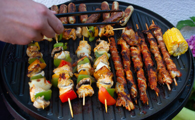 Mans hand is turning a sausage on a home grill on party with barbecue, meat skewers and sausages, bacon, vegetable.