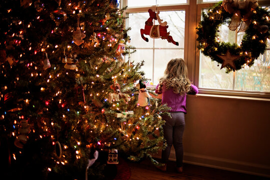 Toddler looking out window next to colorful Christmas tree