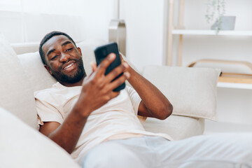 African American man sitting on a black sofa, using his smartphone with a happy expression He is typing a message, enjoying the comfort of his modern apartment The image depicts a young man at home,