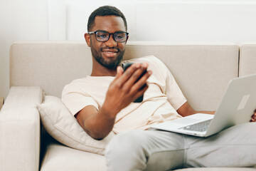 Smiling African American man working on a laptop, sitting comfortably on a sofa in his modern living room He is multitasking, typing on his phone and having a video call while doing online shopping