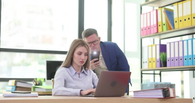Woman types on laptop and man peeps taking photo of screen using phone. Female expresses irritation with unscrupulous behavior of colleague