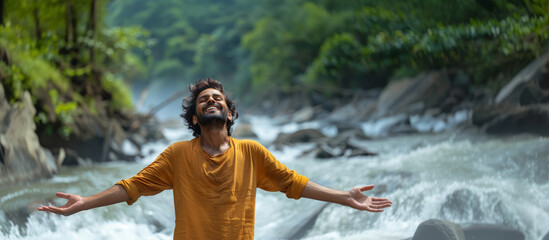 An Indian man standing outdoors with arms spread open with landscape of river or stream in the background. Freedom, nature, inspiration, motivation, relaxation, and independence concept