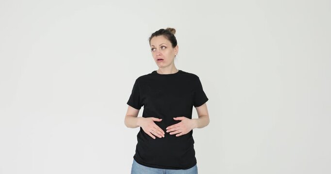 Woman touches stomach letting out heavy sigh on gray background. Displeased lady in black t-shirt feels discomfort from having overeaten in restaurant