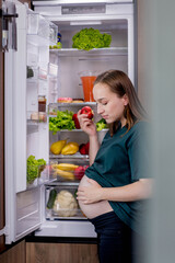 Pregnant woman is eating an apple near the refrigerator