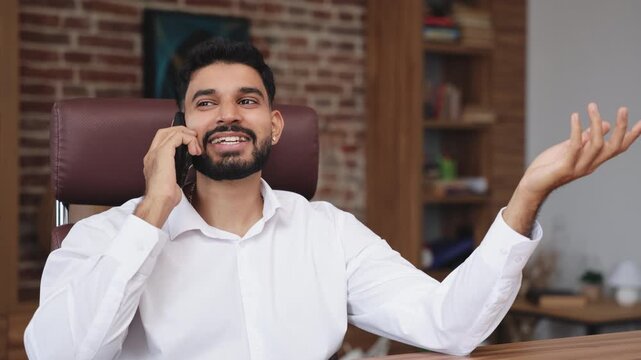 Portrait of talkative indian male having conversation on modern smartphone while gesturing with hands in modern office environment. Smiling man actively explaining something indoors.