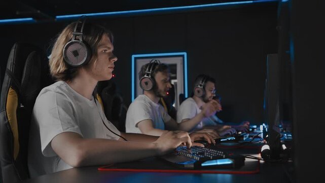 Pro team is playing real-time strategy at cybercafe during tournament. Five players are playing tactical shooter game. Gamers in white T-Shirts are participating in E-sports competition at gaming cafe