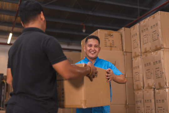 Two men manually carrying a large cardboard board around the warehouse. One smiling directly at camera. Handling newly arrived shipment.