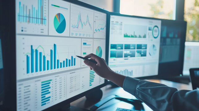 A close-up of a multi-monitor setup displaying various data graphs and charts, with a person pointing at one of the screens. - Powered by Adobe