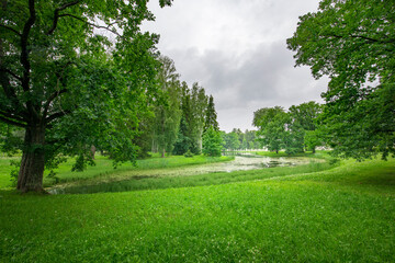 A tranquil park features vibrant green grass and towering trees, while a gentle waterway flows through the lush landscape under overcast skies.