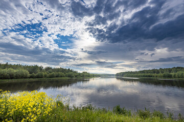 A tranquil river scene with lush greenery along the banks and a bright blue sky partially obscured by puffy white clouds.