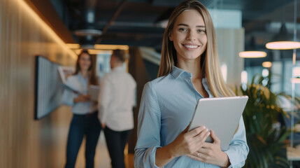 Professional businesswoman confidently holding a tablet, standing in a modern office with colleagues in the background, showcasing leadership and ambition.