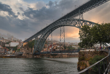 Naklejka premium The iconic Dom Luis I Bridge spans the Douro River in Porto, Portugal, with a warm sunset glow. Traditional hillside buildings and a serene riverbank enhance the scene.