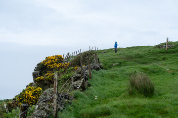 Frau wandert an einem Holzzaun an den Klippen von Nordirland und steht da mit blauer Jacke und...