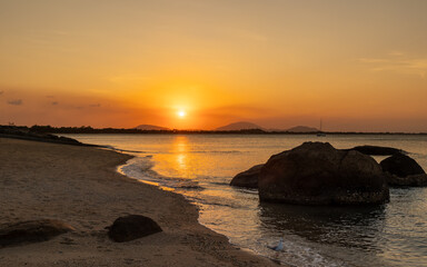 Fototapeta premium An orange sunset with clouds over a calm bay and with silhouetted granite rocks in the water and on the beach in a long exposure photo at Gray's Bay in Bowen in tropical Queensland, Australia.