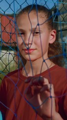 Vertical: Young female football player in sports uniform makes hole in net with hand to better see mates coming to court. Teenage girl with ball under arm gazes into distance on football field closeup