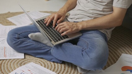 Naklejka premium Focused adult man college student sitting on the floor in apartment room at daylight and doing homework typing on laptop. Education concept.