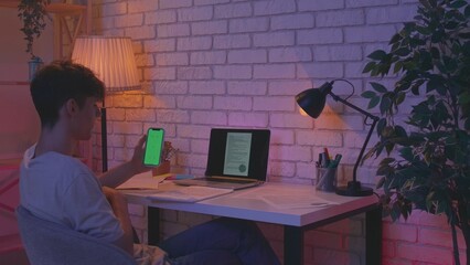 Fototapeta premium Focused adult man college student sitting at desk in apartment room late at night and doing homework using smartphone chroma key green screen mockup. Education concept.