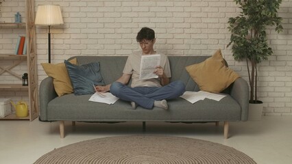 Focused adult man college student sitting on sofa in living room at daylight and doing homework, holding notebook reading essay. Education concept.