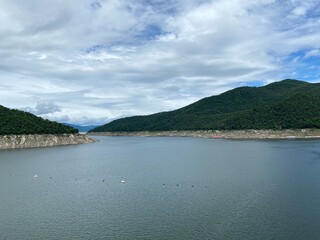 Scenic view of a dam in the mountains with a river, lush green trees, and a clear blue sky at Bhumiphol Dam, Tak, Thailand.
