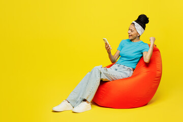 Full body young Latin woman wears blue top t-shirt headband casual clothes sit in bag chair hold in hand use mobile cell phone do winner gesture isolated on plain yellow background. Lifestyle concept.