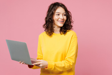 Naklejka premium Young fun happy IT woman she wear yellow sweatshirt casual clothes hold use work on laptop pc computer look aside on area isolated on pastel light pink background studio portrait. Lifestyle concept.