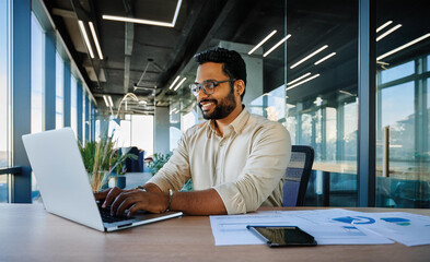 A smiling young man in a suit sits in the office at a desk and works on a laptop, typing on a keyboard, texting, working with data