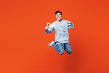 Full body young man wears blue sweatshirt casual clothes jump high hold point on gift certificate coupon voucher card for store isolated on plain red orange color background studio. Lifestyle concept.