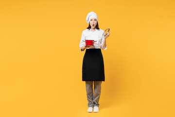 Full body young shocked chef cook baker woman wear white shirt black apron uniform toque chefs hat hold in hand red stewpan lift lid isolated on plain yellow background studio. Cooking food concept.
