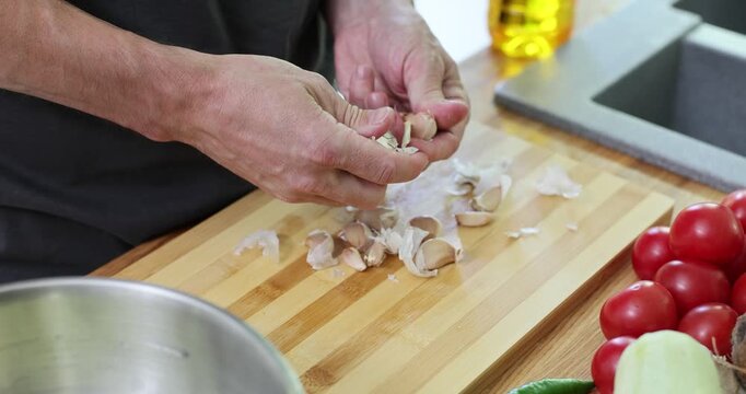 Male hands peel off papery outer layer from garlic clove on cutting board in kitchen. Man prepares organic ingredient to add spicy taste