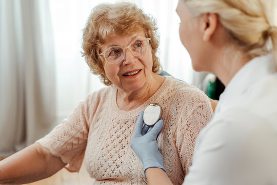 Doctor, cardiologist holding pacemaker, showing to senior patient, sitting at home on sofa