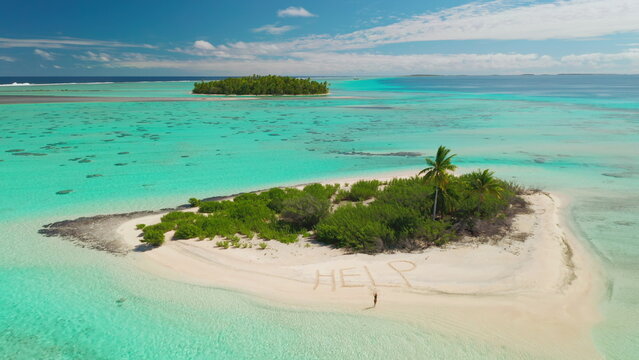Aerial drone footage of a castaway writing help on the beach of a small desert island with palm trees, surrounded by a turquoise lagoon
