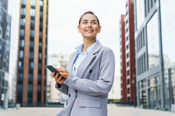 Fototapeta premium Confident businesswoman with a smartphone, standing between modern office buildings, exuding professionalism and success.