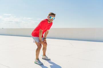 An athletic man in a red shirt and sunglasses takes a break from exercising on a bright, sunny day.