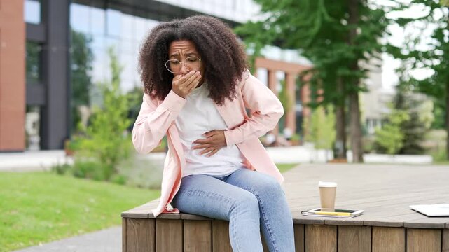 African american female student suffering from nausea, wants to vomit sitting on bench on street near university building. Unhealthy young black woman feels bad, has a stomach ache. She has poisoning