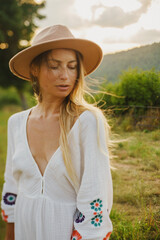 A girl with long blonde hair in a hat, boots and a white long dress on the background of a panorama of green mountains, Alps in Europe at sunset. A girl with a bouquet of lavender flowers, wearing sun