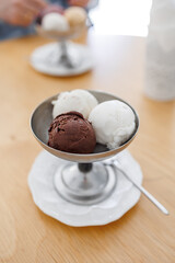 A silver bowl filled with chocolate and vanilla ice cream sits on a white saucer. A silver spoon lays on the table next to the bowl. the dessert is sweet from milk and sugar. summer dessert