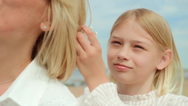 Mother and daughter sitting on picnic blanket on windy beach on weekend. Blonde woman and girl enjoy together leisure and have fun, chatting and laughing. Daughter pins up mother hair and adds flowers