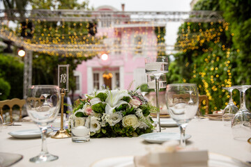 A romantic outdoor wedding reception with round tables, white linens, and floral centerpieces. The tables are set with elegant glassware and silverware, creating a beautiful