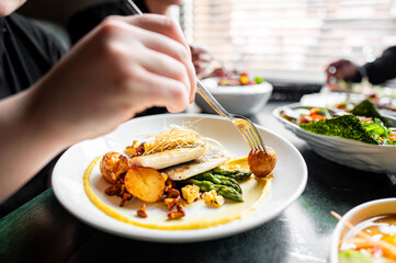 A close-up of hands using utensils to eat a gourmet dish with fish, asparagus, and sliced potatoes on a white plate. The meal is well-presented, healthy, and suitable for food-related content.