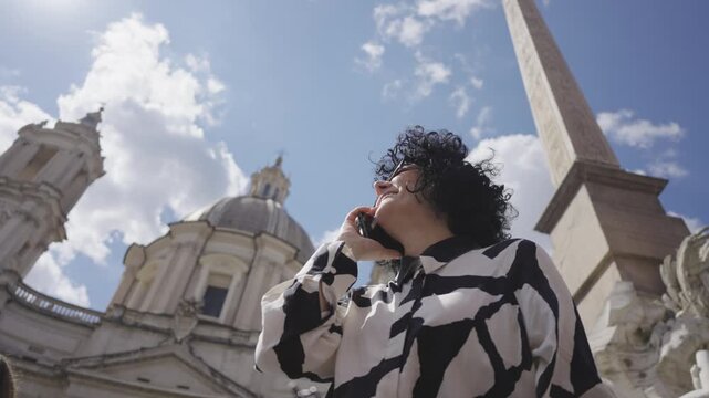 A business woman dressed in a modern black and white blouse talks on her phone, with the historical Sant Agnese church and a striking obelisk in the backdrop under a clear blue sky.