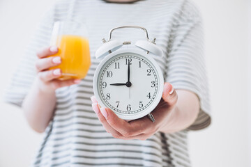 Close-up of a white alarm clock and a glass of orange juice in a female hands.