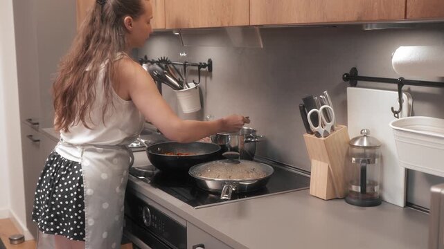 A woman in an apron tastes rice from a pot on the stovetop in her kitchen. She is checking the doneness and flavor of the rice as part of her meal preparation, with pots and pans simmering nearby.