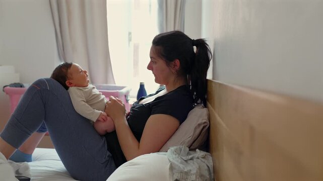 Yawning mother holding her baby, highlighting the fatigue and joy of motherhood. Natural daylight streaming in, creating a warm home atmosphere. Pink bassinet in background