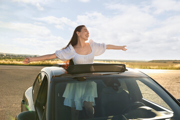 Enjoying trip. Smiling woman leaning out of car roof outdoors